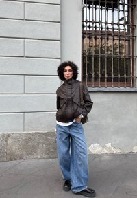 Brązowa skórzana kurtka bombowiec z wysokim kołnierzem, zestawiona z luźnymi niebieskimi dżinsami. Czarne platformy dodają nowoczesnego akcentu. Teksturowane tło.