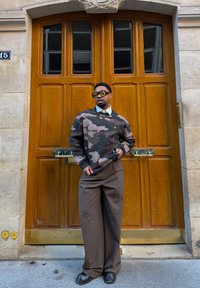 Homme portant un pull camouflage, un pantalon large marron et des lunettes teintées, debout devant des portes doubles en bois sur un bâtiment en pierre.