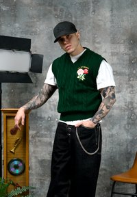 Jeune homme aux bras tatoués portant une casquette noire et un gilet en maille verte, debout à côté d'une vitrine claire en bois contenant des objets musicaux.