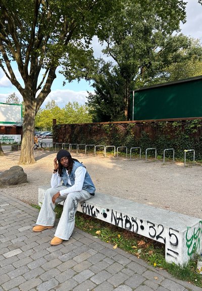 Chaleco de denim sobre una camisa blanca de manga larga, pantalones anchos grises y zapatos slip-on color beige. Sentado en un banco de concreto con graffiti.