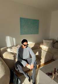 Grauer Strickpullover, dunkle weite Hose, schwarze Sonnenbrille und Lackleder-Pumps. Helles beiges gestreiftes Sofa und blaue abstrakte Kunst im Hintergrund.