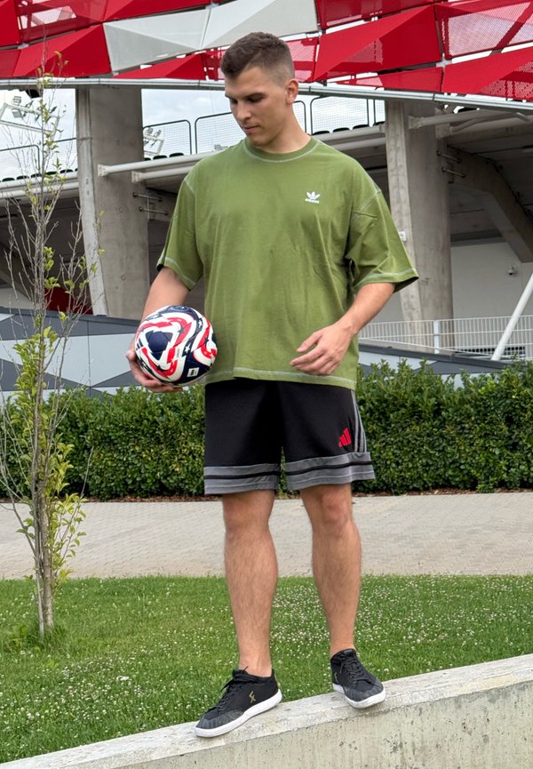 Grønn oversized t-skjorte, svarte shorts med røde detaljer, og svarte treningssko. Holder en multicolor fotball, står på en betongkant.