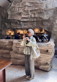 Donna con un foulard beige, occhiali da sole e abbigliamento leggero si trova all'interno di un caffè con un bancone in pietra e un barista che lavora dietro di esso.