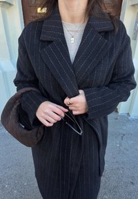 Femme portant un manteau à fines rayures sombres tenant des lunettes de soleil noires et un sac à main tressé marron, avec une bague en or et des colliers superposés visibles.