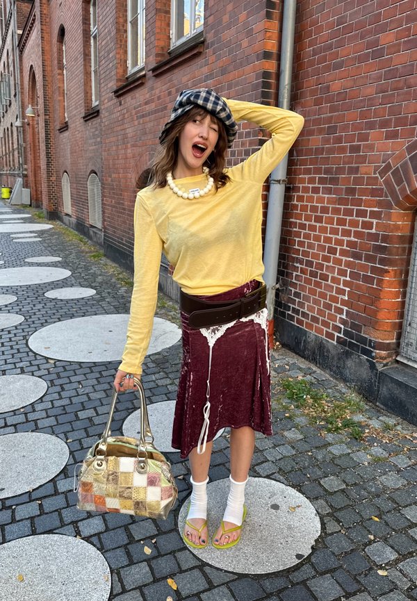 Jonge vrouw in gele top en bordeaux rok poseert op kasseienstraat, houdt een patchwork handtas vast, draagt een geruite hoed en sandalen met sokken.