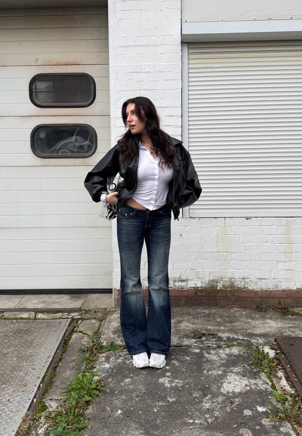 Giacca di pelle nera, camicia bianca a bottoni, jeans a campana blu, sneakers bianche, in piedi contro un muro bianco textured e una porta del garage.