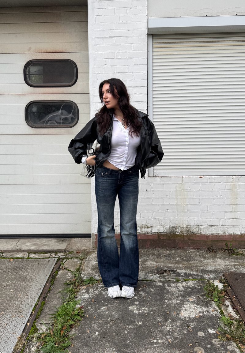 Black leather jacket, white button-up shirt, blue flared jeans, white sneakers, standing against a textured white wall and garage door.