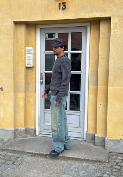 Suéter de rayas verticales grises, jeans de pierna ancha azul descolorido y zapatos negros. De pie cerca de una puerta blanca con paneles de vidrio y una pared amarilla.