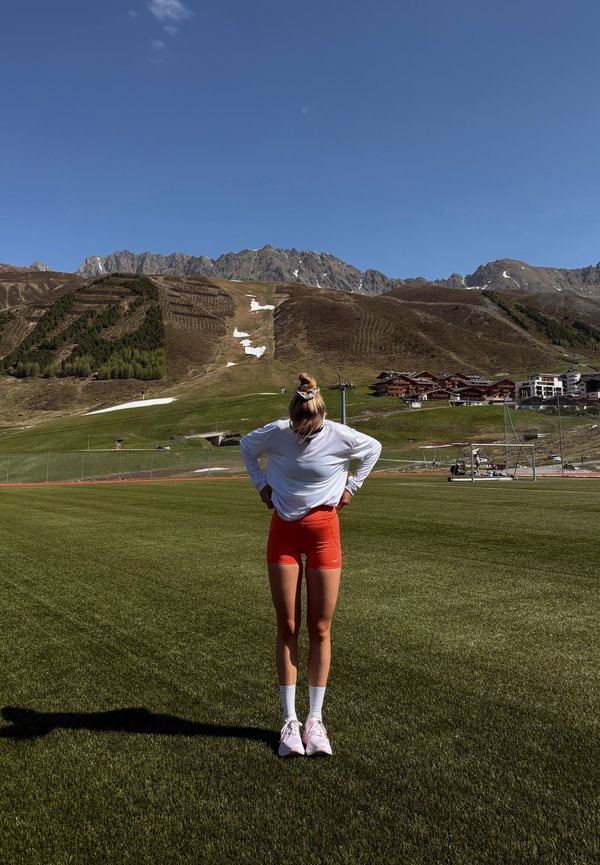 Junge Frau in roten Shorts und weißem Top steht auf einem grünen Sportplatz mit Bergen und Gebäuden im Hintergrund unter klarem blauem Himmel.