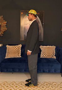 Νεαρός άνδρας με σακάκι ριγέ, γκρι παντελόνι, κίτρινο καπέλο και μαύρα αθλητικά παπούτσια στέκεται πλάγια σε ένα σαλόνι με μπλε καναπέ και αφηρημένη τέχνη.