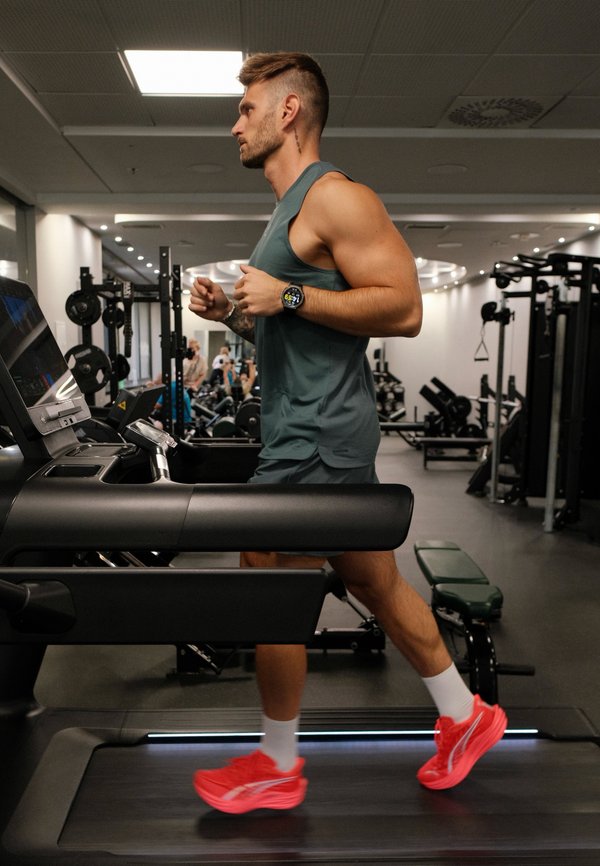 Un hombre corre en una cinta de correr en un gimnasio. Lleva un reloj deportivo, ropa de entrenamiento oscura, calcetines blancos y zapatillas de color rojo intenso.