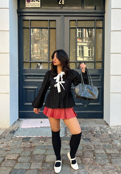 Blusa peplum negra con un lazo blanco, falda roja plisada, medias altas negras hasta la rodilla y zapatos blancos. Sosteniendo un bolso negro; de pie cerca de una puerta.