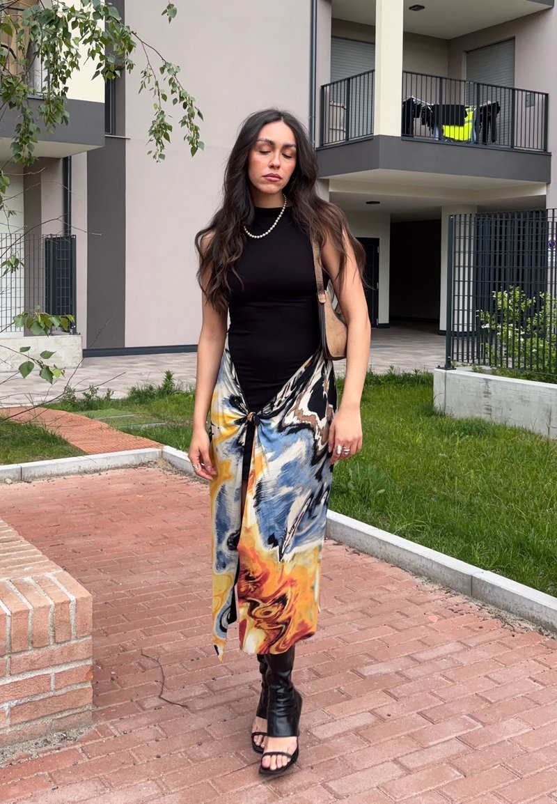 Femme aux longs cheveux foncés portant un haut noir sans manches, une jupe colorée à motifs, des sandales à talons noires et un collier de perles, debout à l'extérieur.