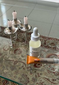 Διαφανές γυάλινο τραπέζι με τρία ροζ κεριά σε ασημένιες βάσεις, ένα λευκό μπουκάλι σταγόνας με την ένδειξη "The Ordinary" και μία πορτοκαλί και γκρι σωλήνα L'Oréal.