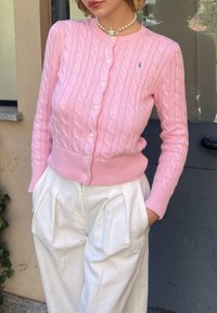 Ανοιχτό ροζ πουλόβερ με πλέξη καλωδίου και κουμπιά στο μπροστινό μέρος, με ριμπ μανσέτα. Φοριέται πάνω από λευκά πλισέ παντελόνια. Διακριτική λεπτομέρεια λογότυπου στο στήθος.