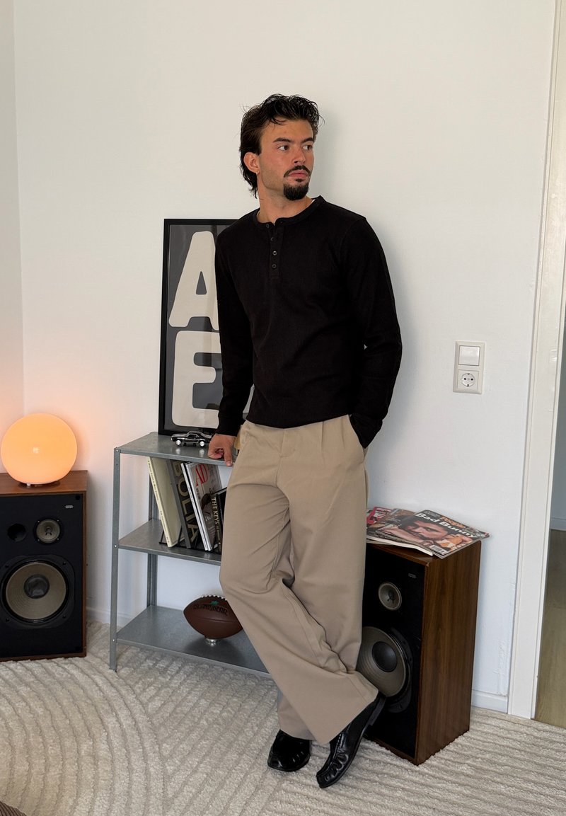 Homme avec une chemise noire et un pantalon beige appuyé contre une enceinte dans une pièce moderne avec des livres, une lampe et des lettres encadrées sur une étagère.