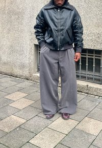 Czarna skórzana kurtka typu bomber z kieszeniami na zamki błyskawiczne, w połączeniu z szarymi szerokimi spodniami. Obuwie w bordowym kolorze. Teksturowane tło.