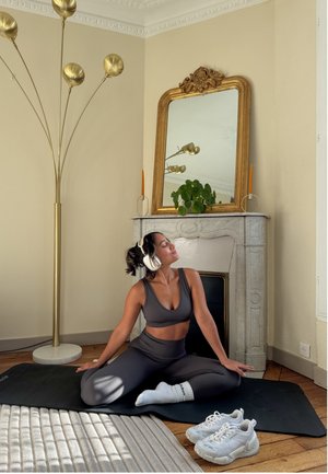 Mujer con ropa deportiva y auriculares sentada con las piernas cruzadas sobre una esterilla en una habitación con un espejo decorativo, una planta y una lámpara de pie cerca.