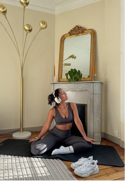 Mujer con ropa deportiva y auriculares sentada con las piernas cruzadas sobre una esterilla en una habitación con un espejo decorativo, una planta y una lámpara de pie cerca.