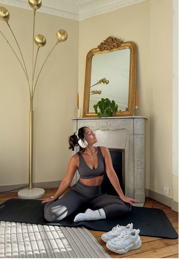 Kvinne i treningsklær og hodetelefoner sitter med beina i kors på en matte i et rom med et dekorativt speil, plante og gulvlampe i nærheten.