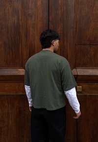 Homme portant des chemises superposées, debout face à une grande porte en bois avec un trou de serrure à hauteur de la taille.