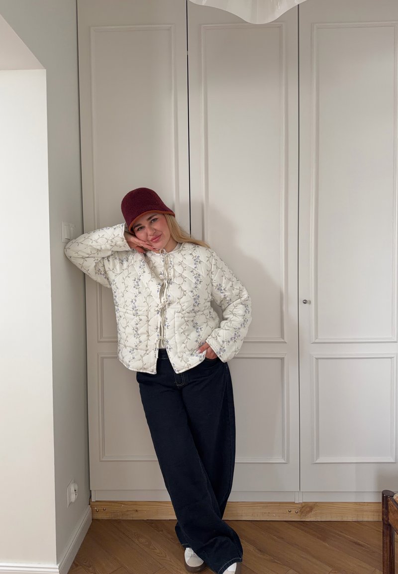 Giovane donna con giacca trapuntata bianca a fiori, pantaloni larghi scuri, scarpe bianche e cappello secchiello color amaranto, appoggiata a una parete bianca a pannelli.