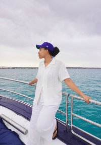 Modell som bär en vit, kortärmad meshtröja, vita byxor och en lila keps, stående på ett båtens reling över turkos vatten.