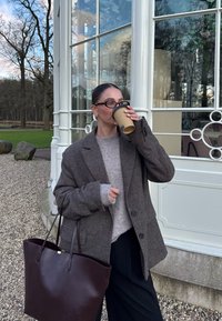 Een bruin wollen blazer over een grijze gebreide sweater, gecombineerd met een zwarte broek. Houdt een koffiekop vast en draagt een bruine leren tote bag.