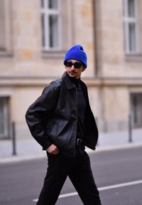 Μαύρο δερμάτινο μπουφάν με γιακά, φορεμένο πάνω από μαύρο λαιμόκο επιστημονικό. Μπλε πλεκτή σκούφια καιoversized μαύρα γυαλιά ήλιου. Λεπτό μαύρο παντελόνι.