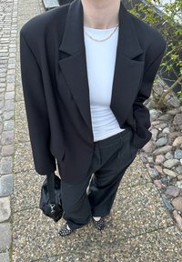 Zwarte oversized blazer over een witte top, gecombineerd met donkere broeken en schoenen met stippen. Een zwarte tas hangt over de schouder.