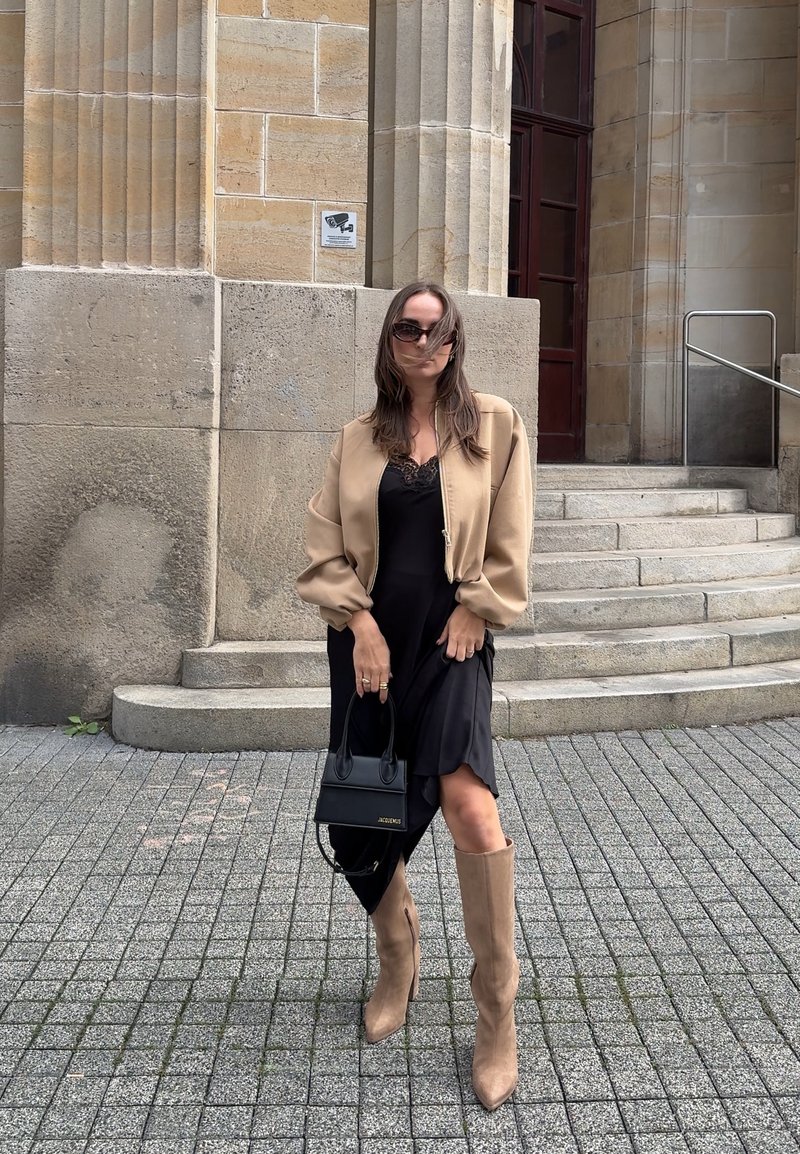 Casaco bomber bege, vestido preto com rendas, botas castanhas até ao joelho e uma pequena mala preta. Texturas suaves e design contemporâneo.
