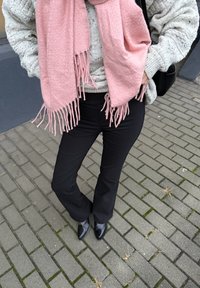 Fekete bővülő szárú nadrág, világosszürke kötött pulóver és rózsaszín rojtos sál. A kifutó a tégla burkolatú járdán áll, fekete magas sarkú lakkcipővel kiegészítve.