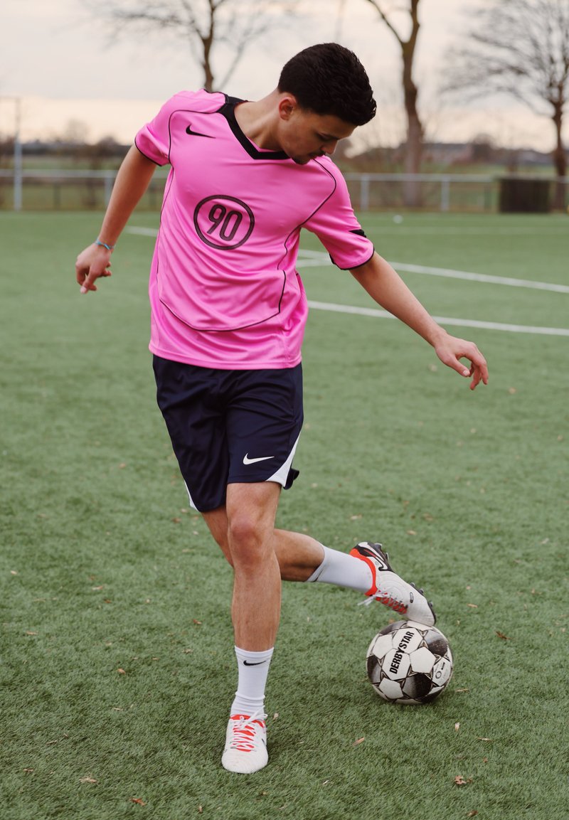 Jeune joueur de football en maillot rose et short noir contrôlant un ballon noir et blanc sur un terrain extérieur vert.