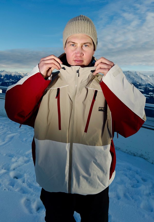 Person iført beige og rød vinterjakke og beige strikket lue som glatter opp kragen i fjellandskap med snø under en blå himmel.