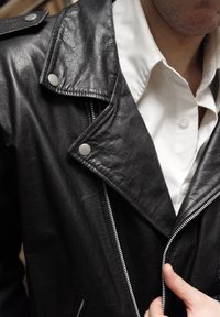 Veste en cuir noir avec fermetures argentées, dotée d'un col cranté, d'une fermeture éclair et de deux poches frontales. Portée par-dessus une chemise blanche.