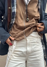 Personne portant un jean blanc taille haute, une chemise marron à fermeture éclair, une veste sombre avec doublure à carreaux, une bague rouge et un collier pendentif en pierre. Les mains ajustent les vêtements.