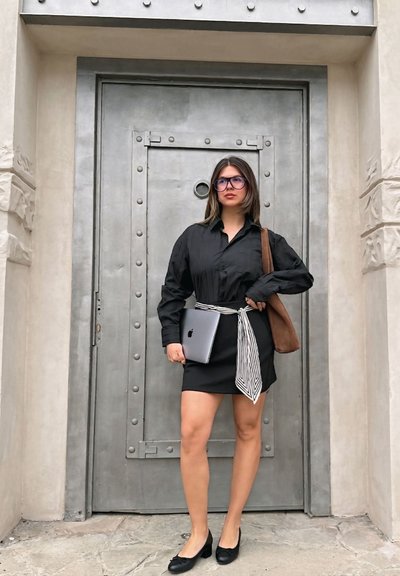 Mujer con vestido negro y cinturón a rayas, sosteniendo un portátil, de pie frente a una gran puerta metálica con paredes de piedra.