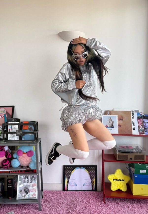 Jeune personne portant une veste argentée, une jupe à sequins, des chaussettes blanches jusqu’aux genoux et des lunettes de soleil, sautant à l’intérieur près d’étagères avec des livres et des peluches.
