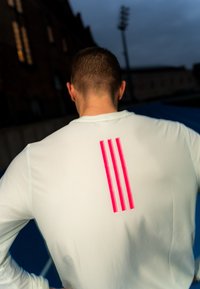 Homme aux cheveux courts portant une chemise blanche à manches longues avec trois bandes verticales roses sur le haut du dos, debout à l'extérieur au crépuscule.