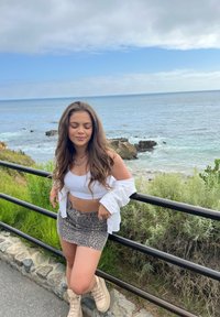 White long-sleeve shirt, white cropped tank top, patterned gray mini skirt, and beige boots; standing by a railing with ocean and greenery in background.