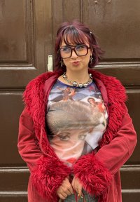 Piros kordbársony kabát műszőrme gallérral, stilizált nőket ábrázoló grafikai póló, ezüstös vastag nyaklánc, kockás als�órész. Barna háttér.