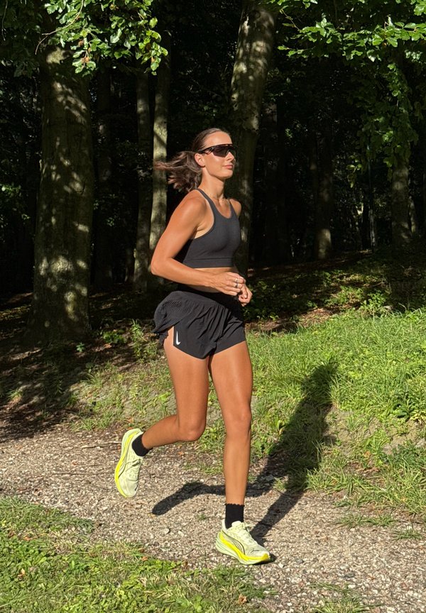 Mujer con ropa deportiva negra y gafas de sol corriendo por un sendero forestal con luz solar y árboles de fondo.