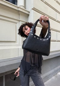 Schwarze Lederhandtasche mit strukturiertem Finish, geschwungener Form, oberem Griff und Metallnieten entlang der Öffnung. Fokussiert auf die Frontansicht.