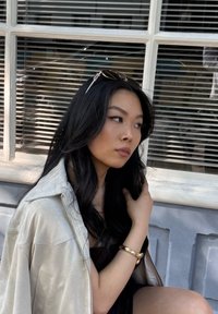 Una giacca leggera di un beige pallido con una texture lucida, indossata sopra un top scuro; presenta un bracciale dorato sul polso sinistro e occhiali da sole nei capelli.