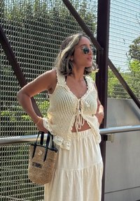 Crochet Top in crema con bordi in pizzo, legato davanti, abbinato a una gonna bianca fluida. Tiene una piccola borsa intrecciata con la scritta "ST TROPEZ".