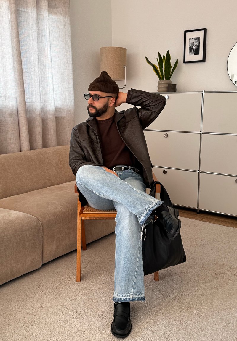 Άνδρας με καφέ σκουφί, γυαλιά ηλίου, δερμάτινο μπουφάν και ανοιχτόχρωμο τζιν κάθεται σταυροπόδι σε ξύλινη καρέκλα σε μπεζ σαλονάκι.