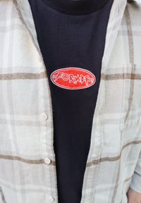 Chemise en flanelle à carreaux blancs et beiges portée sur un t-shirt noir ornée d'un patch ovale rouge avec le mot « FORMER » en lettres blanches.