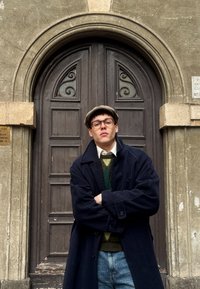 Une personne se tient devant une porte en bois, portant un manteau bleu foncé, un pull à motifs verts et jaunes, des lunettes et une casquette plate grise.