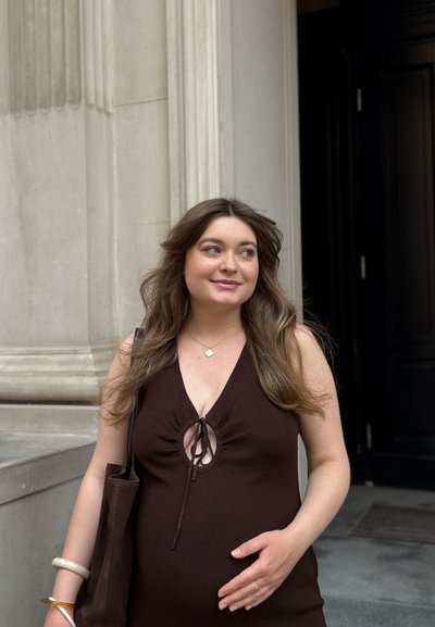 Vestido marrón acanalado con una apertura en forma de llave, escote redondo y lazo ajustable. Tonalidad de piel clara, con una mano sobre su vientre redondeado, sosteniendo un bolso.