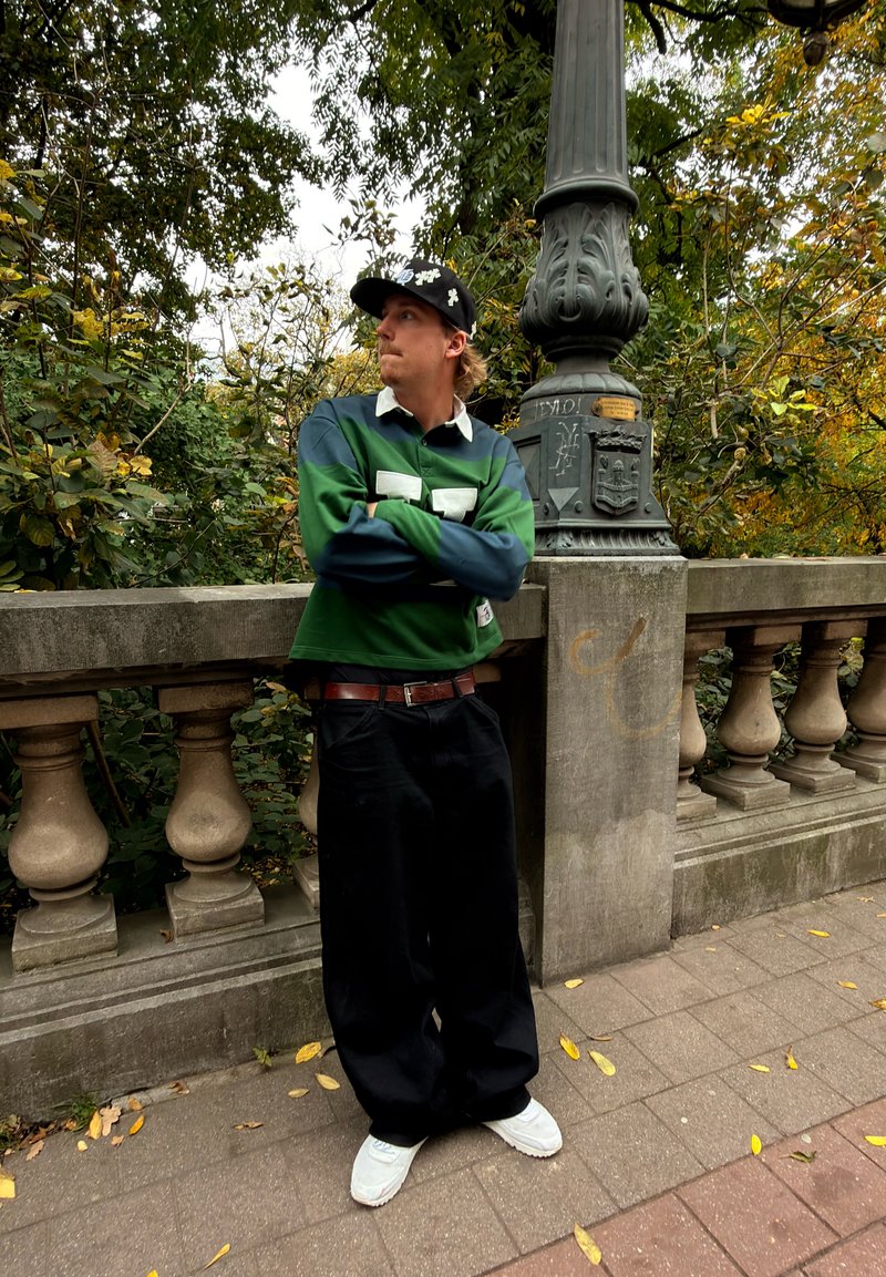 Polo à manches longues vert et bleu avec col blanc, pantalon noir large et chaussures blanches ; debout près d'une rambarde en pierre au milieu du feuillage.
